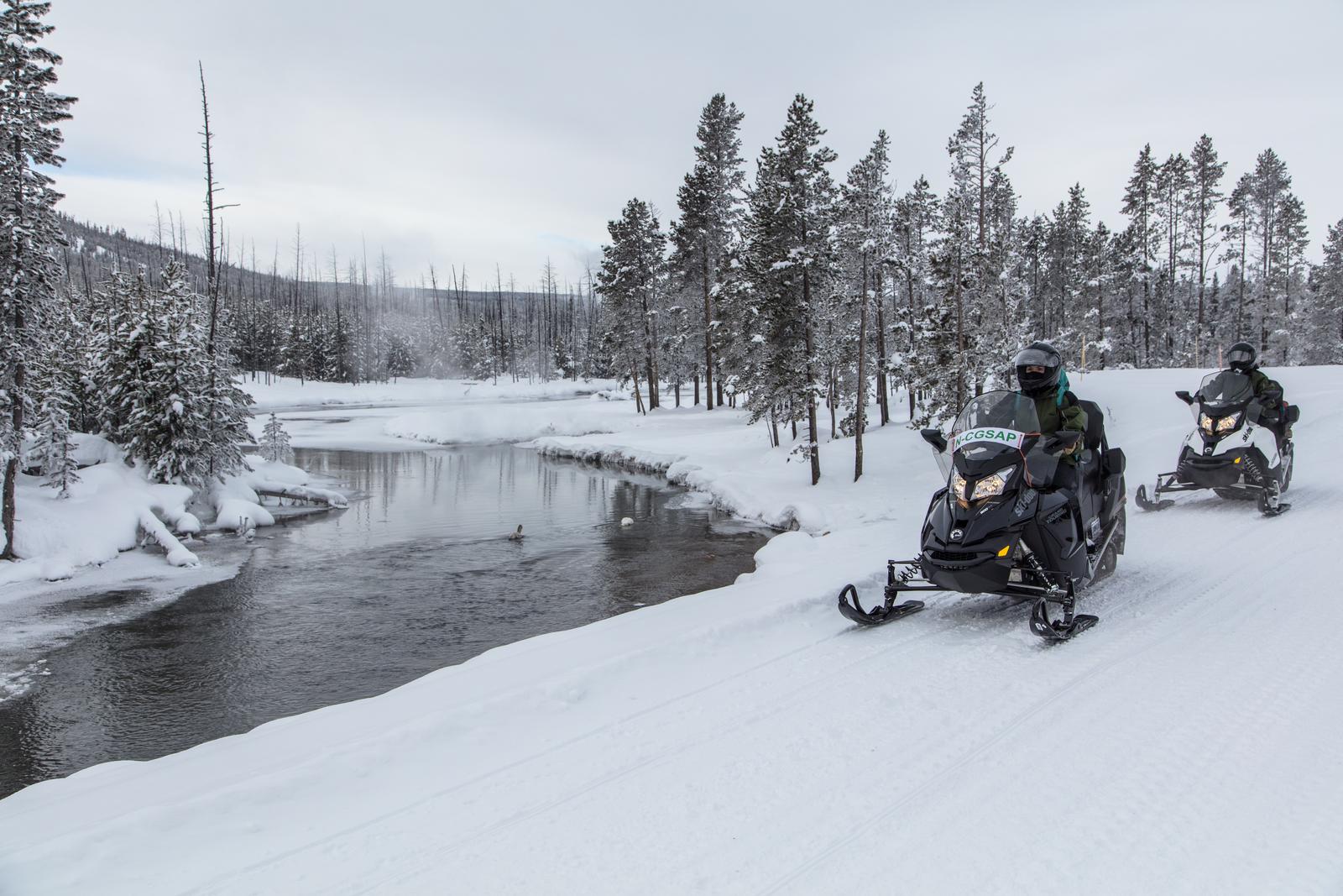 Two snowmobiles travel on the road beside a riverSnowmobiling along the Gibbon River
