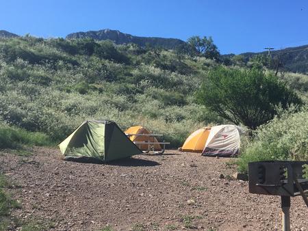 Three tents set up in flat, gravelly area with green mountains in backgroundTent area and grill