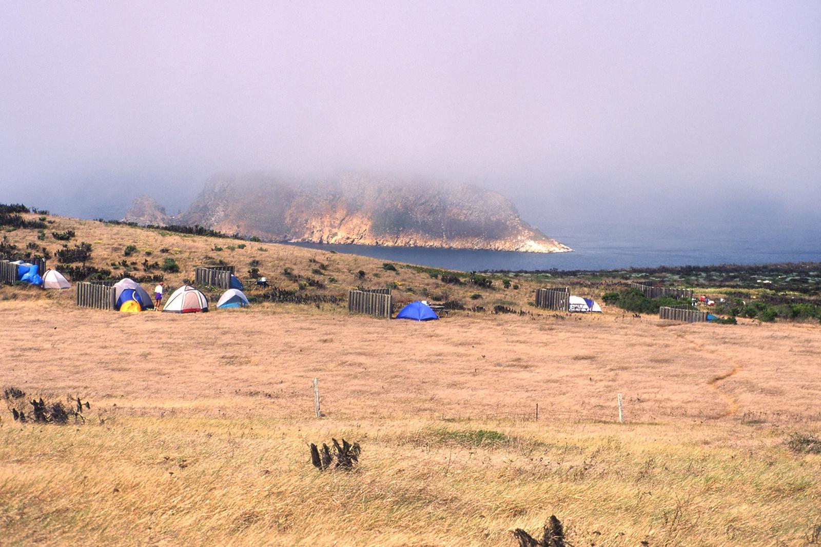 Tents and 5-foot tall windbreaks perches on an ocean bluff overlooking an islet covered in fog. SAN MIGUEL ISLAND
