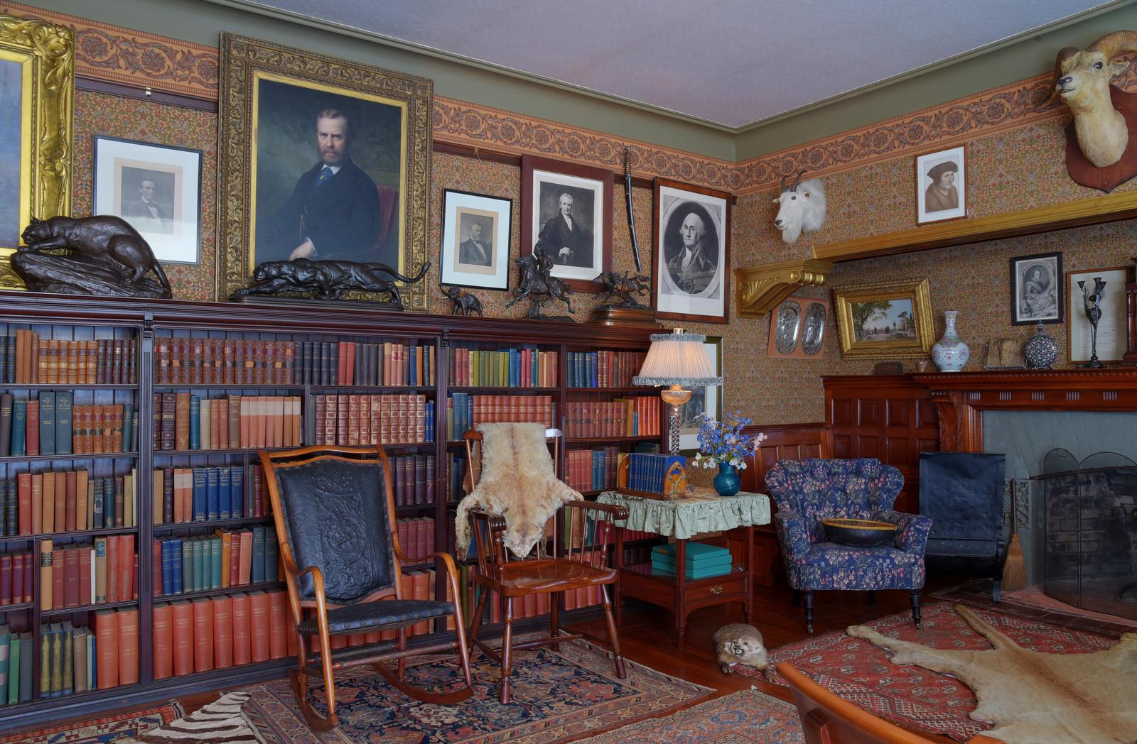 A room with books, paintings, hunting trophies, chairs, and a fireplace.The library.