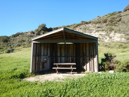 Eight foot tall wind shelter with picnic table surrounded by green grass. Sites 001-015 - 012
