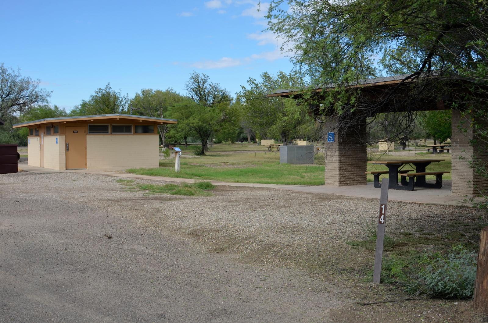 View of ADA accessible Site 14 from the main roadway. There is a wide gravel driveway for parking, a large shade shelter over a picnic table, and a paved sidewalk leading to a restroom. Cottonwood trees and bear boxes from other camp sites can be seen scattered in a grassy field behind the restroom.View of ADA accessible Site 14 from the main roadway.