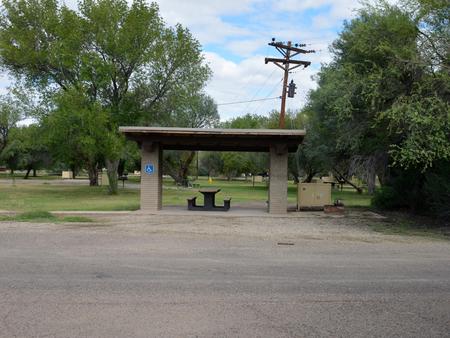 Wide view of the shade shelter, picnic table, and bear box for Site #14. The shelter, picnic table, and bear box are built on a paved area to provide ease of access to the site's amenities. The gravel driveway is directly in front of the shade shelter, allowing the shortest distance possible between a parked vehicle and the campsite. A grassy field with cottonwood trees and a telephone pole can be seen just behind the site.Shade shelter and bear box for Site #14.