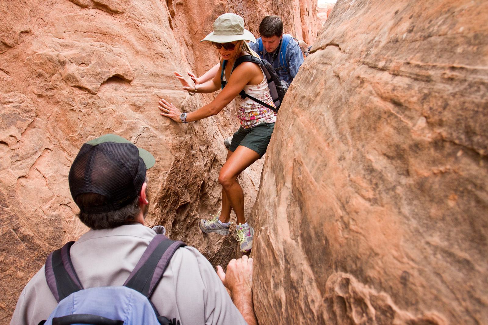 two people moving through a narrow crack between two rocks while a ranger monitorsIn some places in the Fiery Furnace, you must hold yourself off the ground by pushing against the sandstone walls with your hands and feet.
