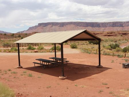 At the Upper Onion Creek Group Site a, two picnic tables sit underneath a large shade shelter with a fire ring  nearby and a horse corral in the distance. Wide open views of the Fisher Valley area surround the site. In the distance are large, red rock plateaus.