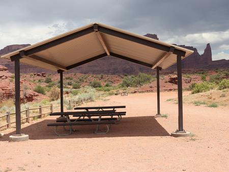 The Upper Onion Creek Group Site B has a large shade shelter with picnic tables underneath, surrounded by undulating, shrub-covered desert land. In the distance, the Fisher Towers area can be seen.