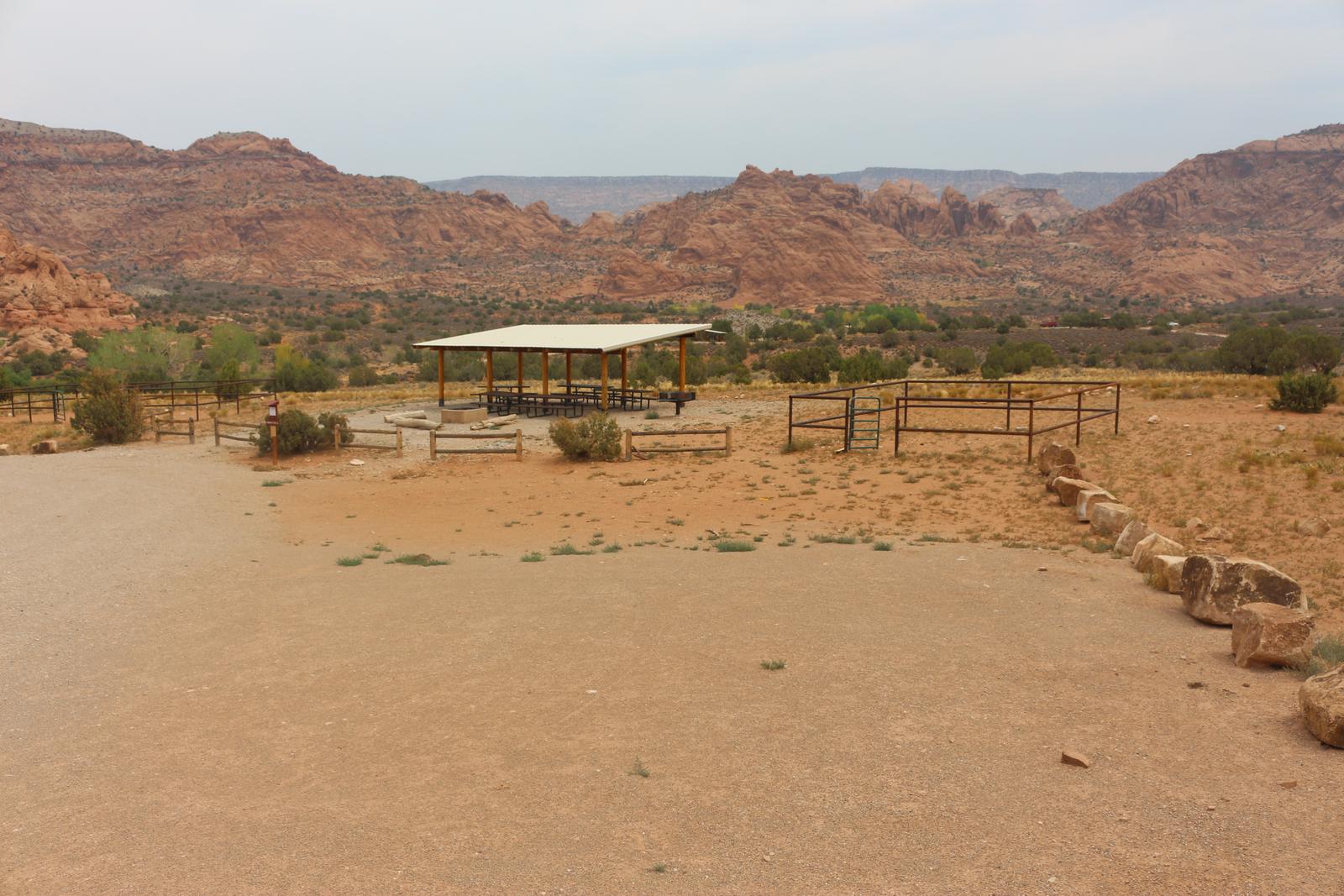 Ken's Lake Group Site A shade shelter, metal fence horse corral, and parking area with undulating, red, slick rock hills on the horizon. 