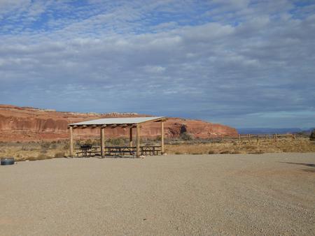 Lone Mesa Group Site A shade shelter, picnic tables, fire pit and parking area. Red rock cliffs in the distance.