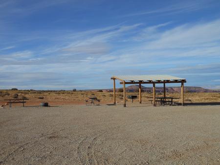 Lone Mesa Group Site C shade shelter, picnic tables, fire ring, and parking area. Flat, open desert lands in the distance.
