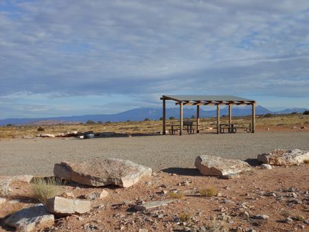 Lone Mesa Group Site C shade shelter, picnic tables, parking area, and fire pit with the La Sal Mountains in the distance.