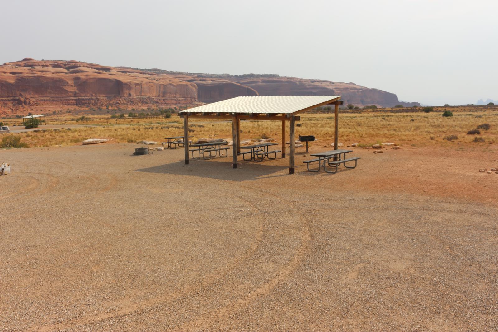 Lone Mesa Group Site E shade shelter, picnic tables, fire ring, and standing grill. Tall, red rock plateaus line the horizon.