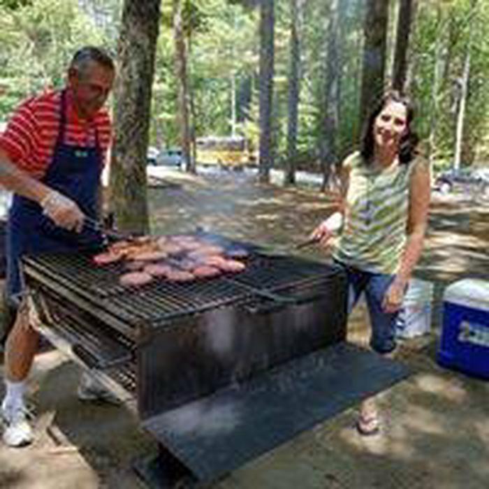 Two adults, man in apron, cooking burgers on a charcoal grill at the shelter area.  Coolers in background and forested area.Serve it your way with family and friends.  Grills provided at all shelters and picnic groves.