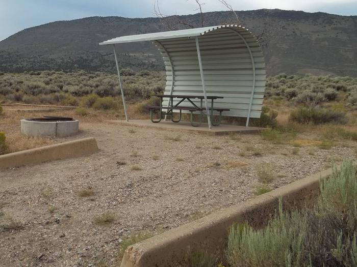 Picnic table with shelter on one side and overhead. There is a fire pit with a grill grate in front of the picnic table and a tent pad off to the side.Antelope Campground: Site 6