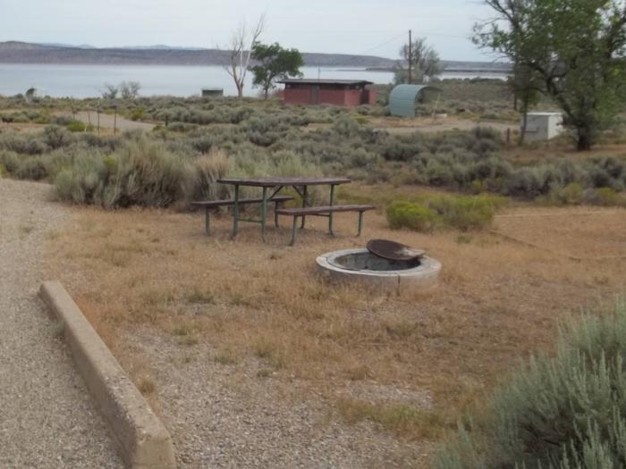 Near the picnic table, you will find a fire pit that has a grill grate. There is a tent pad lined with timbers off to the side and a red brick build that houses the restrooms is in the background.Antelope Campground: Site 9
