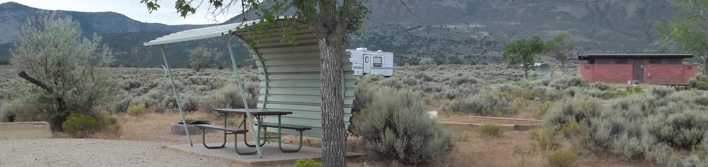 Partial sheltered picnic table with a timber border tent pad near by.  A red brick building is located in the background that houses the restrooms.Antelope Flat Campground: Site 26