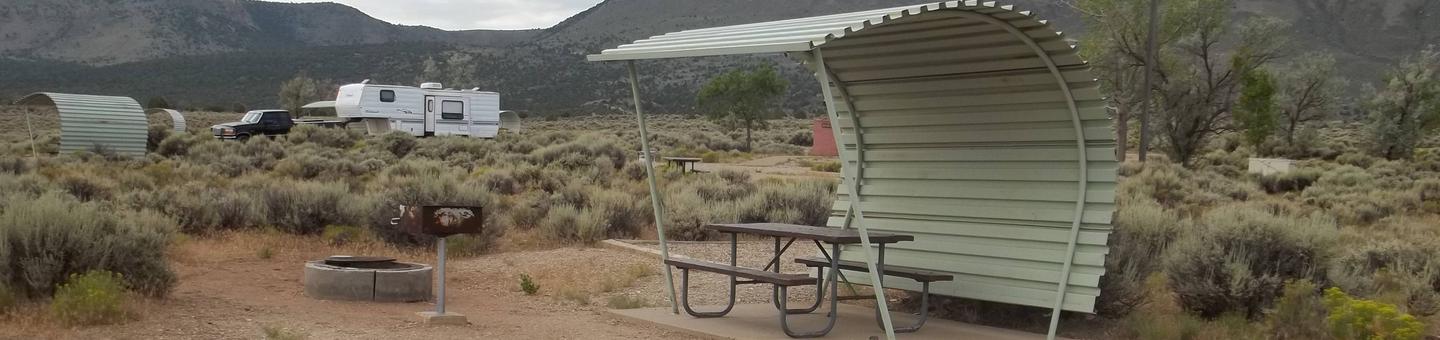 Partial covered picnic table near a grill and fire pit. Campers are in the background in another site.Antelope Flat Campground: Site 28