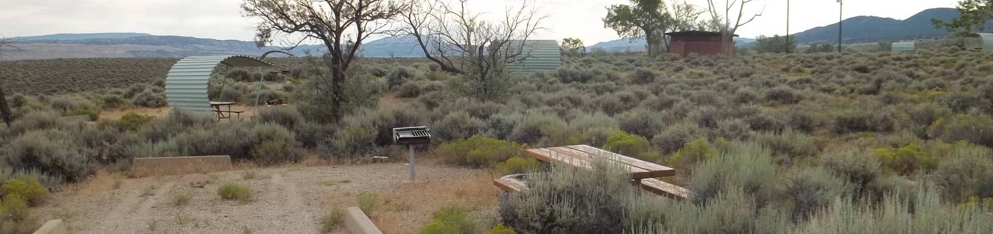 Picnic table with grill in the campsite surrounded by sagebrush. Another site and red building with restrooms is in the background.Antelope Flat Campground: Site 35