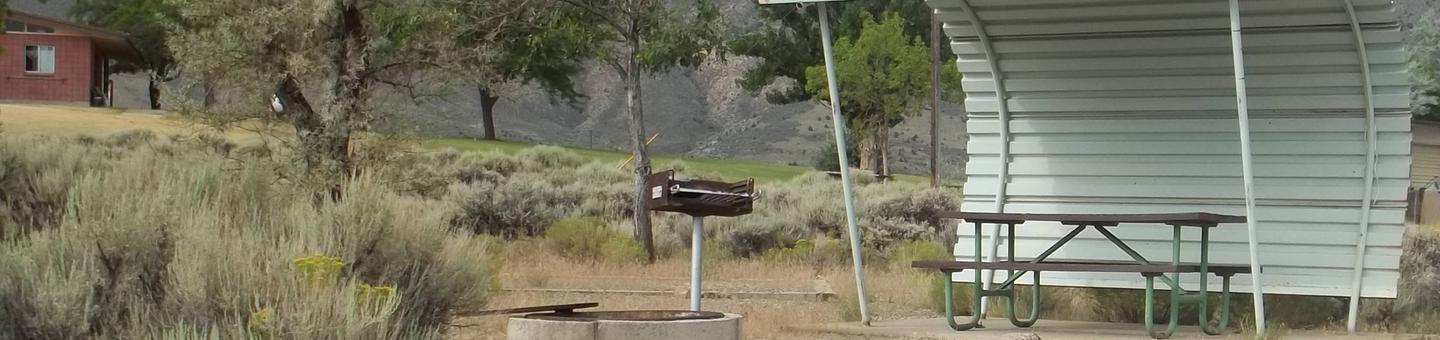 Partial covered picnic table with grill and fire pit on the side. A red building that houses the restrooms in the in the background.Antelope Flat Campground: Site 36