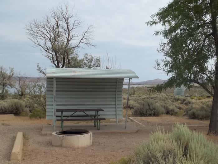 Partial covered picnic table with a fire pit in front of it and a tent pad off to the side. The lake can be seen in the background.Antelope Flat Campground: Site 39
