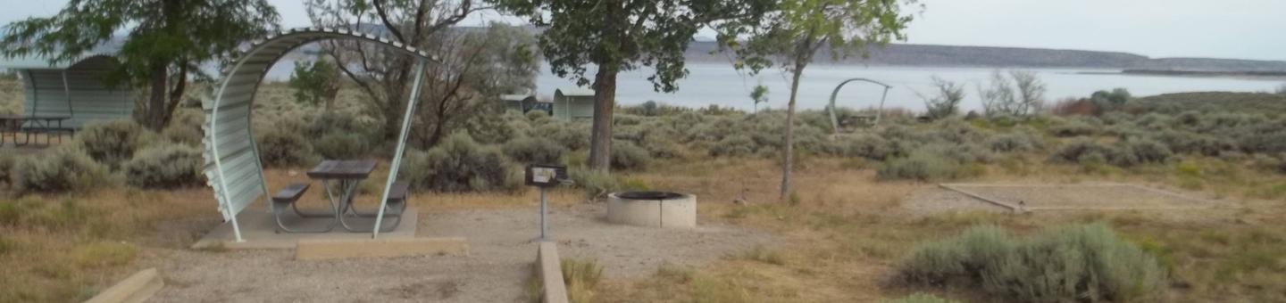 Partial covered picnic table with a grill, fire pit, and tent off to the side. A lake is in the background.Antelope Flat Campground: Site 40