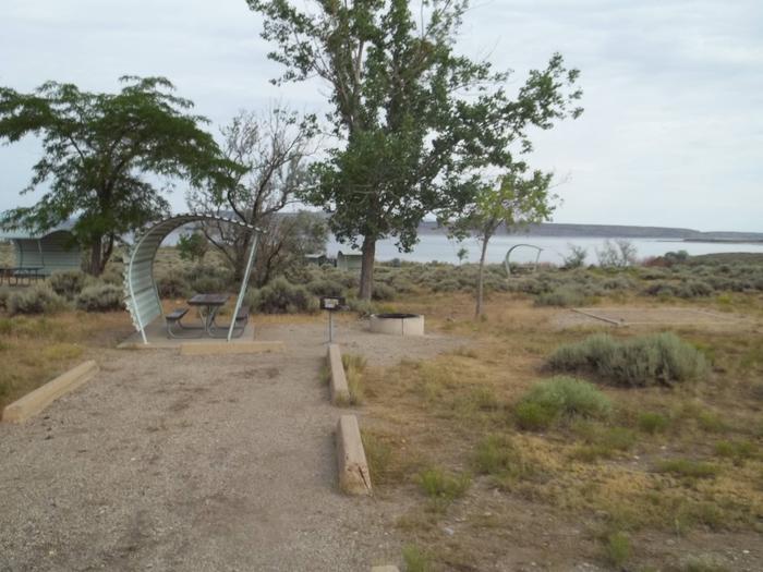 Partial covered picnic table with a grill, fire pit, and tent off to the side. A lake and another site is in the background.Antelope Flat Campground: Site 40