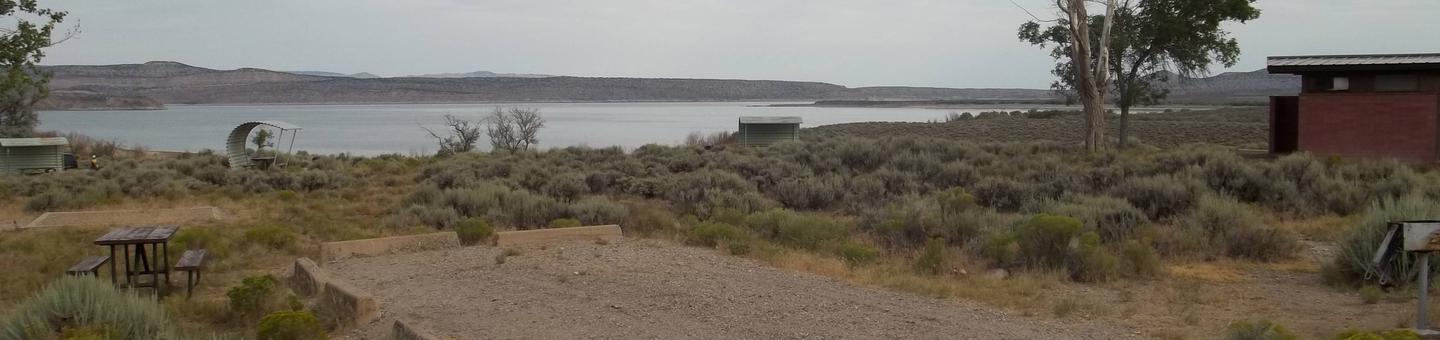 Picnic table on the side of parking strip with a grill on the other side. The lake and building with restrooms is in the background.Antelope Flat Campground: Site 41