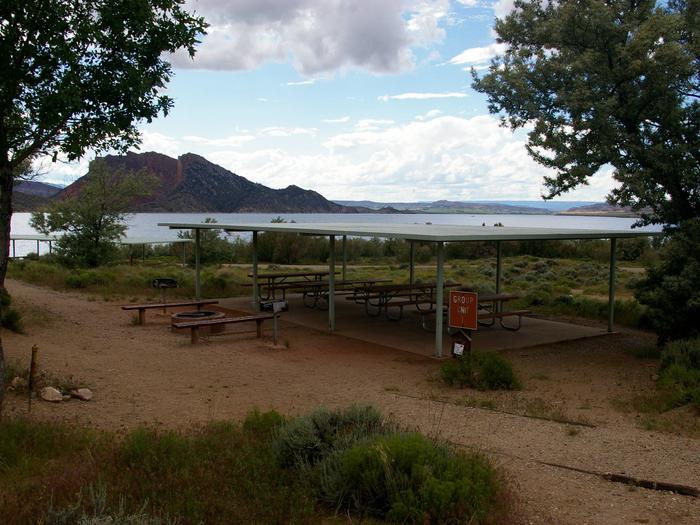 Pavillion that covers a number of picnic tables and a fire pit with some seating around it sets off to the side. The lake can be seen in the background.Antelope Flat Campground - Group Site 1