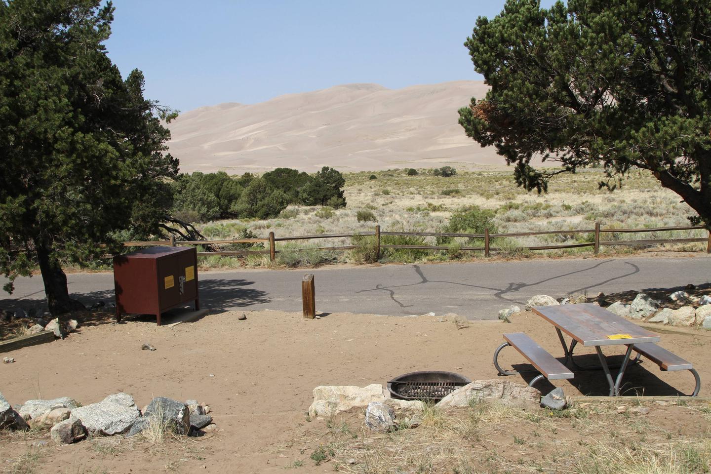 Back view of Site #24 tent pad, picnic table, fire ring, and bear box. Sand Dunes can be seen in the background.Site #24, Pinon Flats Campground