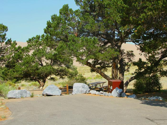 View of Site #26 parking pad, fenced and designated tent pad, bear box, and picnic table. Parking pad has medium boulders around the pavement.Site #26, Pinon Flats Campground