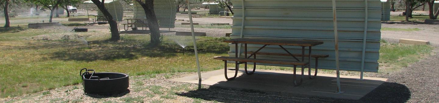 Partial covered picnic table on a slab of cement and a fire pit  with a grill grate off to the side in a semi grassy area.Buckboard Crossing Campground: Loop A, Site 11