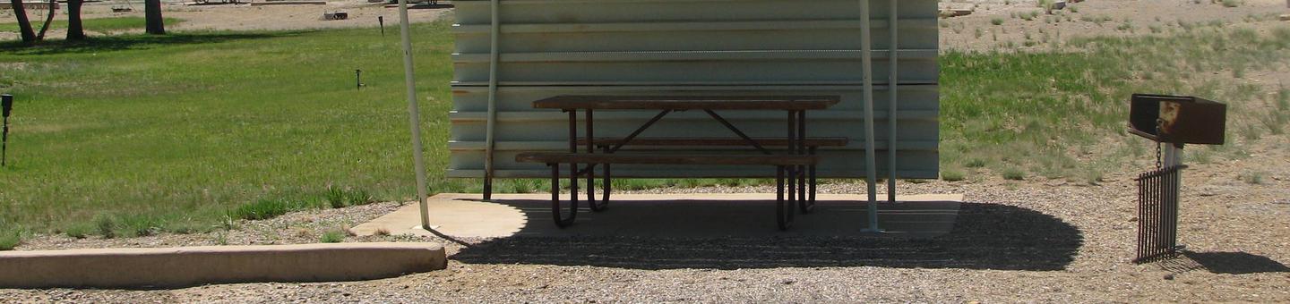 Partially covered picnic table on a slab of cement with a grill and grate off to the side in the gravel.Buckboard Crossing Campground: Loop B, Site 12