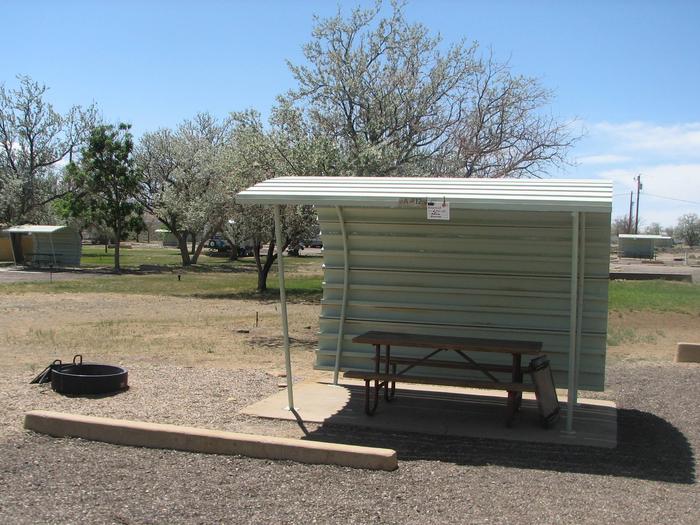 Partically covered picnic area on a slab of cement and a fire pit with grate off to the side in the gravel.  Other campsites are in the background.Buckboard Crossing Campground: Loop A, Site 12