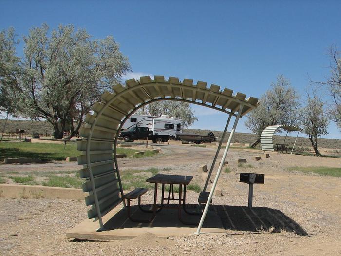 Partially covered picnic table on a slab of cement with a grill off to the side in the gravel. There is a truck and RV trail in the background at another site.Buckboard Crossing Campground: Loop B, Site 13