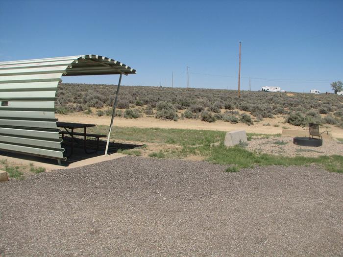 Partially covered picnic table on a slab of cement. A fire pit and grill slates is found to the side of the picnic area in the gravel. Sage-brush flats with utility poles are in the background.Buckboard Crossing Campsite: Loop A, Site 15
