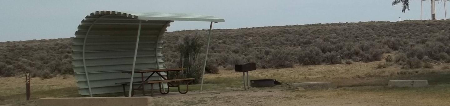 Partially covered picnic table on a slab of cement. There is a grill near the table. The background is filled with sagebrush.Buckboard Crossing Campground: Loop B, Site 16