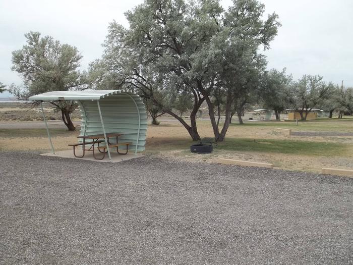 Partially covered picnic table on a slab of cement with a fire pit nearby. The restrooms are located in the yellow building in the background.Buckboard Crossing Campground: Loop A, Site 17