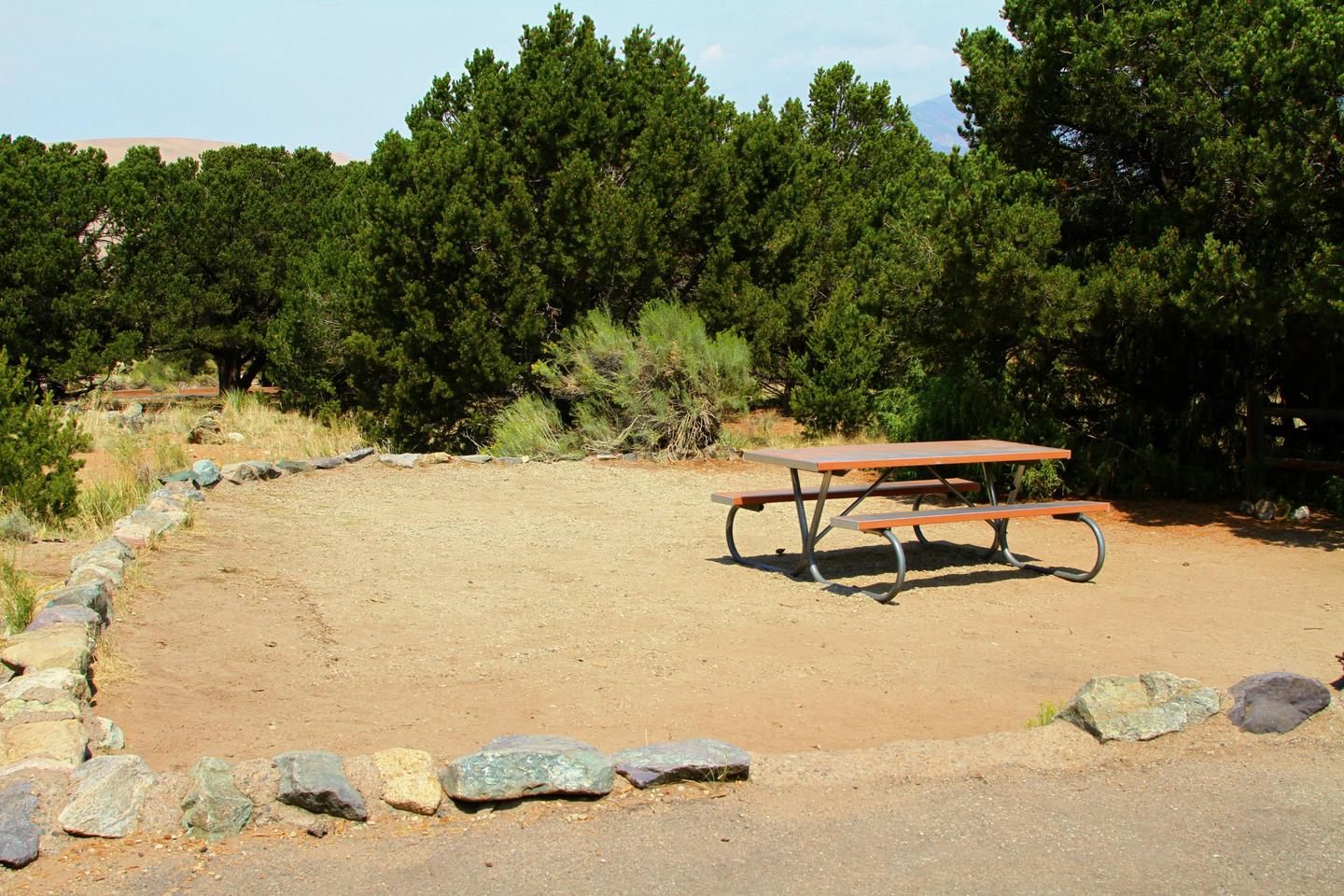 Closer view of Site #57 tent pad with picnic table. Site has pine trees on one side of boundary and is ringed by a low rock border.Site #57, Pinon Flats Campground