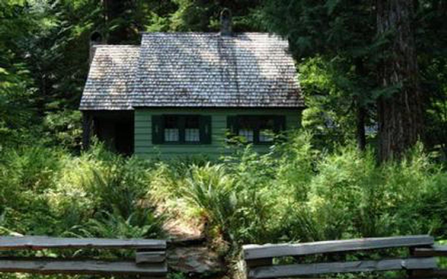 GOVT MINERAL SPRINGS GUARD STACabin with shake roof and green clapboard siding surrounded by ferns and conifer trees.
