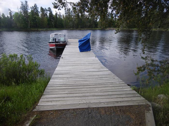 R26 - Sunrise Point, view looking from campsite of the boat dock with a boat tied to it and accessibility swing with blue cover.View looking out from boat access