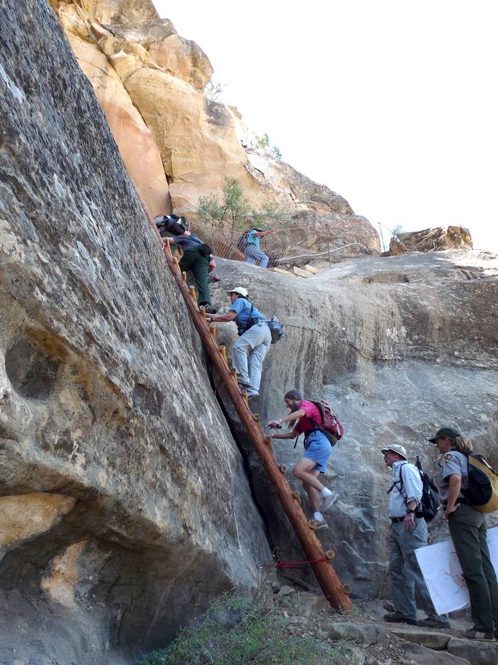 People climbing ladder and stone steps along a steep cliff faceVisitors and ranger exiting Square Tower House.