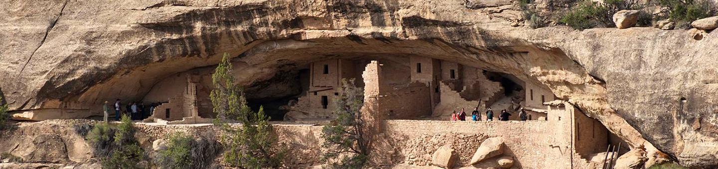 People exploring an ancient stone masonry village in a cliff alcoveVisitors on a ranger-guided tour of Balcony House.