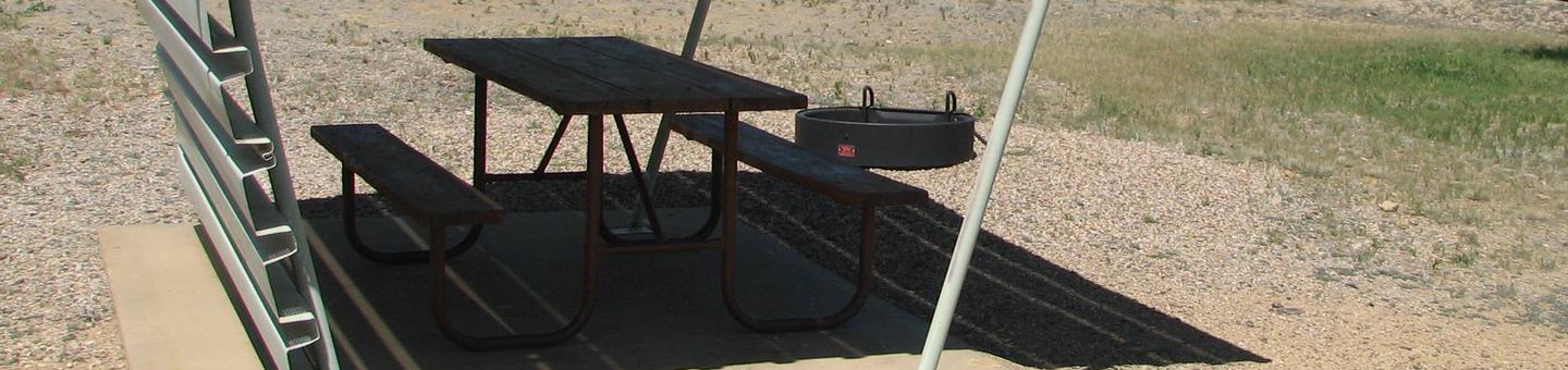 Partially covered picnic table on a slab of cement with a fire pit in the gravel off to the side.Buckboard Crossing Campground: Loop A, Site 19