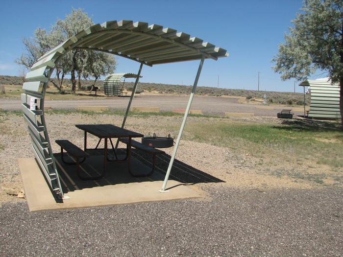 Partially covered picnic table on a slab of cement with a fire pit in the gravel off to the side.Buckboard Crossing Campground: Loop A, Site 19
