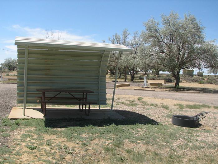 This site has a partially covered picnic table on a slab of cement and a fire pit with a grill grate nearby that is in the gravel.Buckboard Crossing Campground: Loop A, Site 21