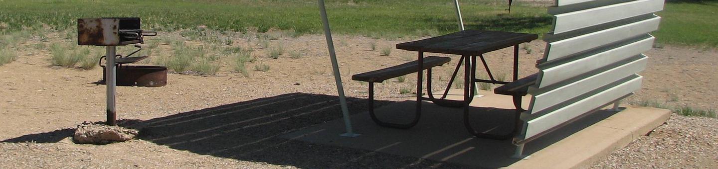 This site has a partially covered picnic table on a slab of cement with a grill next to it held up by a metal rod in the ground and a fire pit found in the gravel.Buckboard Crossing Campground: Loop B, Site 21