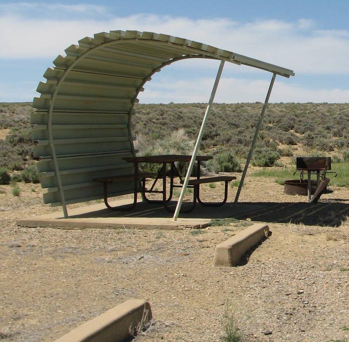 This site has a partially covered picnic table on a slab of cement with a grill next to it held up by a metal rod in the ground and a fire pit found in the gravel.Buckboard Crossing Campground: Loop B, Site 22