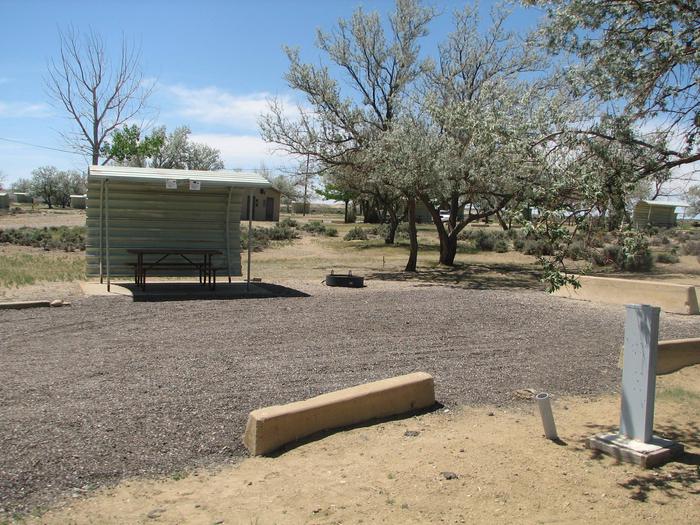 This site has a partially covered picnic table on a slab of cement and a fire pit found in the gravel. The restroom facilities are in the background.Buckboard Crossing Campground: Loop A, Site 22