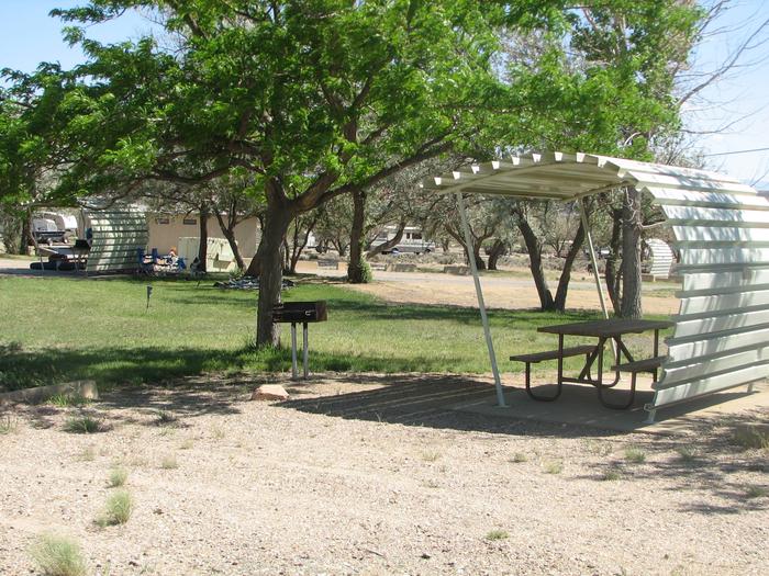 This site has a partially covered picnic table on a slab of cement with a grill next to it held up by a metal rod in the ground.Buckboard Crossing Campground: Loop B, Site 23