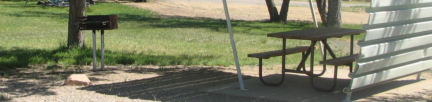 This site has a partially covered picnic table on a slab of cement with a grill next to it held up by a metal rod in the ground.Buckboard Crossing Campground: Loop B, Site 23