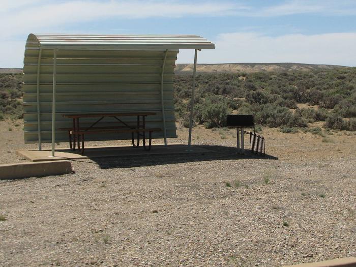 This site has a partially covered picnic table on a slab of cement with a grill next to it held up by a metal rod in the ground.Buckboard Crossing Campground: Loop B, Site 24